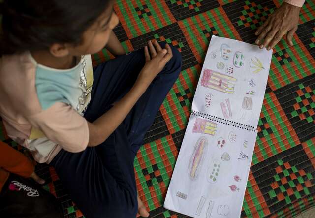Ein Kind sitzt auf einem gemusterten Boden und betrachtet Zeichnungen oder Notizen auf einem Blatt Papier, während eine weitere Hand darauf zeigt.